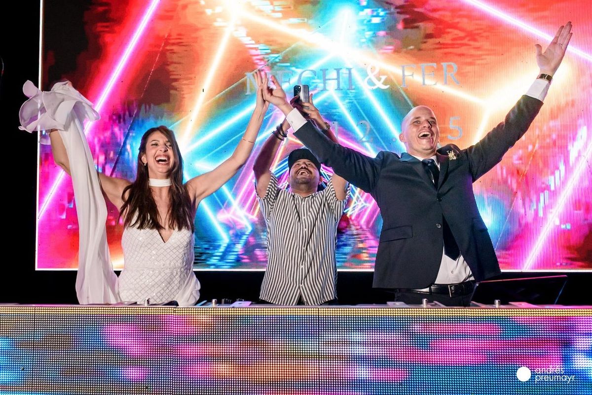 Los novios en la cabina de DJ con los brazos en alto y pantalla LED de fondo